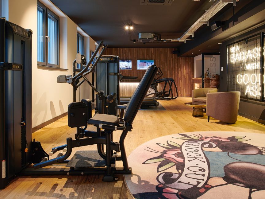 Fitnessraum mit schwarzen Kraftgeräten (Matrix), Holzboden, rundem Teppich, Neonschrift und Loungesessel.