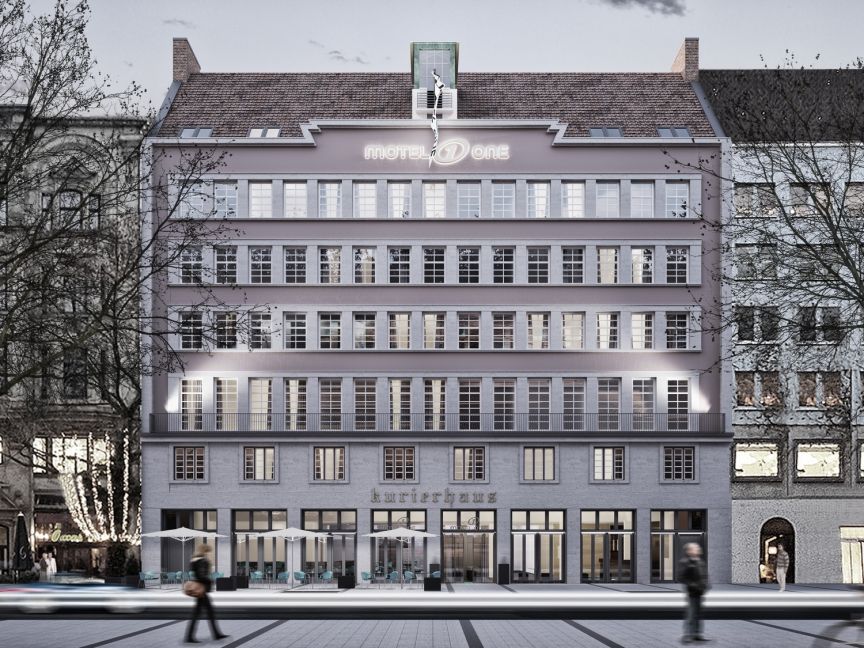 Mehrstöckiges Stadtgebäude „Motel One Kurierhaus" bei Dämmerung, beleuchtet, mit Passanten und kahlen Bäumen.