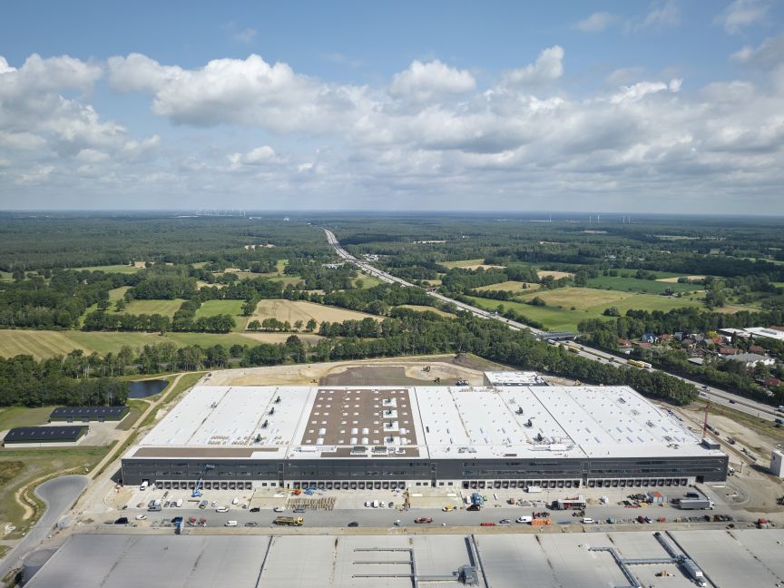 Luftaufnahme eines großen Lagergebäudes mit Laderampen, umgeben von Autobahn, Feldern und Wäldern.