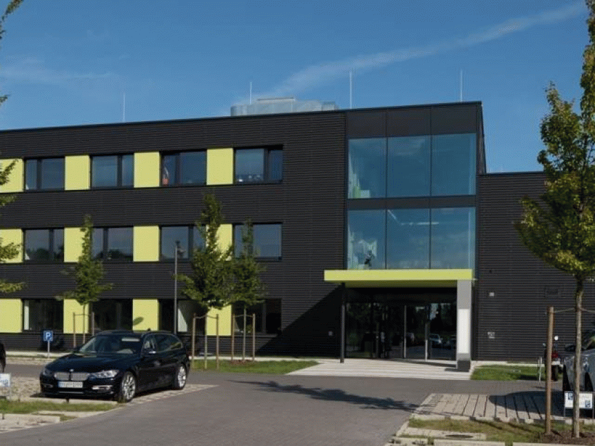 Dreigeschossiges Bürogebäude mit dunkler Fassade und gelben Akzenten, davor Parkplatz mit Autos und Bäumen.