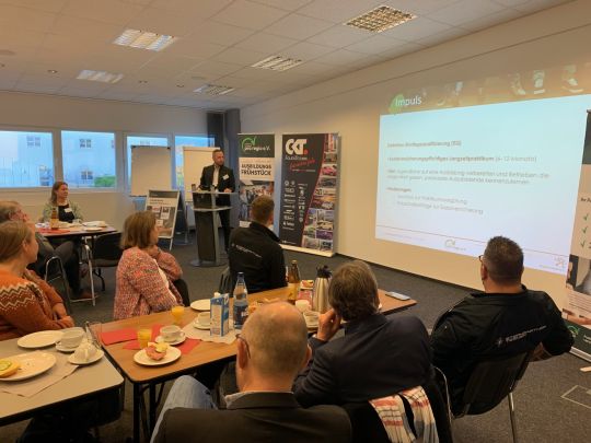 Mehrere Personen sitzen am Frühstückstisch und bekommen parallel eine Präsentation von einem Referenten vorgestellt. 