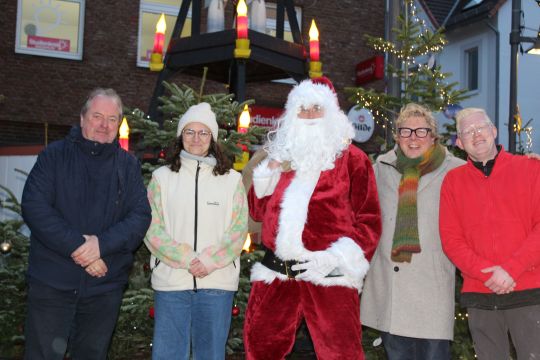 Fünf Personen stehen vor einem Weihnachtsbaum, wobei die Person in der Mitte wie der Weihnachtsmann verkleidet ist.