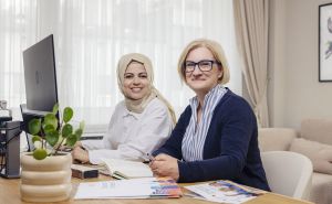 Zwei Frauen sitzen an einem Schreibtisch mit Computer, Dokumenten und Pflanze; eine trägt Hijab, die andere Brille.