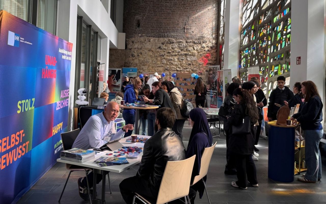 Messestand mit Besucher:innen in einer Halle; buntes Banner mit deutschen Wörtern wie „Stolz", „Happy", „Selbstbewusst".