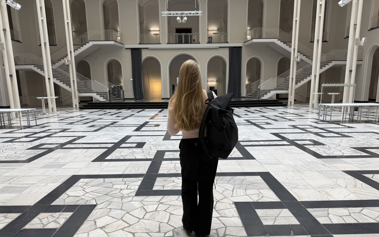 Frau mit langen blonden Haaren und schwarzem Rucksack steht in einer großen, hellen Halle mit gemustertem Marmorboden.