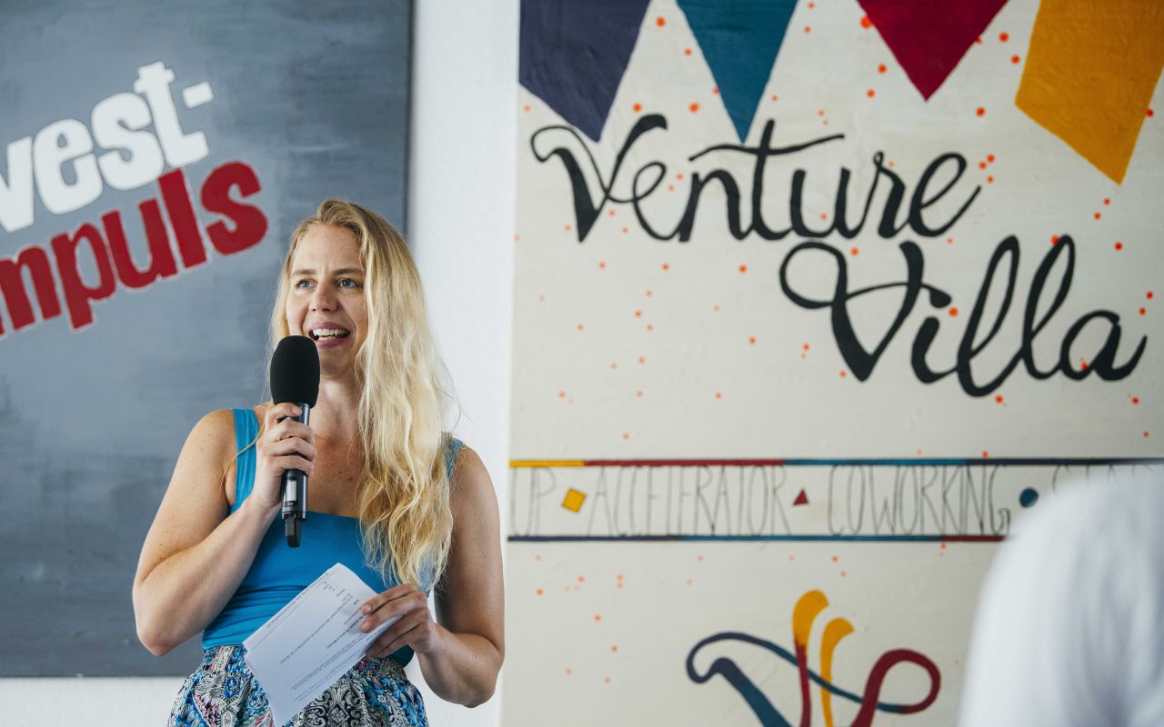Blonde Frau mit Mikrofon spricht vor einem Venture Villa Schild, hält Papier in der Hand.