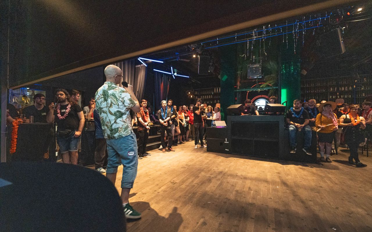 Person in Hawaiihemd steht auf Bühne vor Publikum in dunklem Club mit Holzboden und Neonbeleuchtung.
