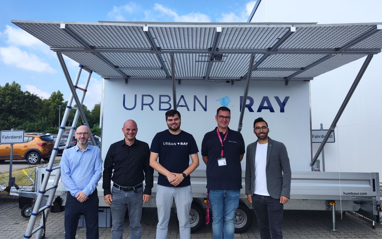 Fünf Männer stehen vor einem weißen Anhänger mit Solarpanelen und dem Logo „Urban Ray" bei bewölktem Himmel.