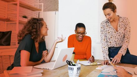 Drei Frauen sitzen und stehen an einem Tisch mit Laptop und Notizen und unterhalten sich lächelnd.