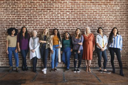 „Zehn Frauen unterschiedlichen Alters und Herkunft stehen lächelnd vor einer Backsteinwand."