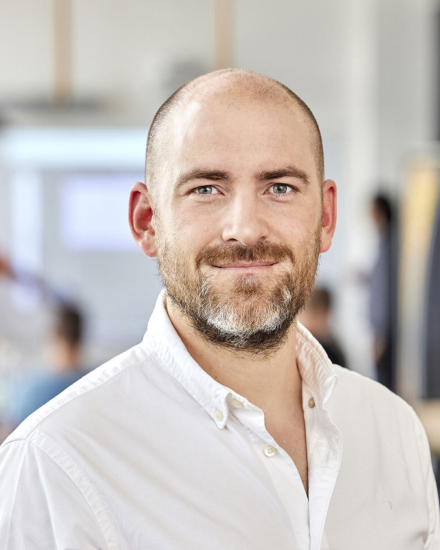 Mann mit Glatze und Bart, blaue Augen, weißes Hemd, lächelt in die Kamera, unscharfer Büro-Hintergrund.