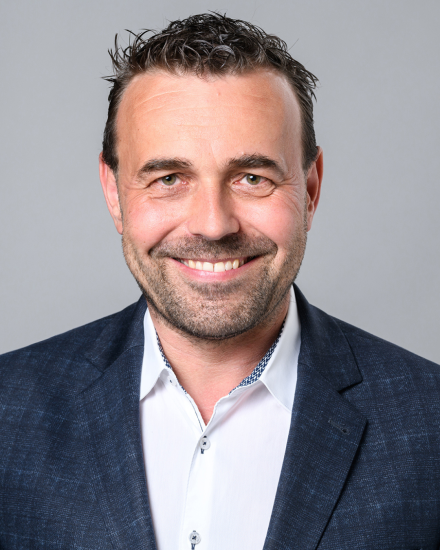 Mann mit dunklem Haar und Bart, weißem Hemd und blauem Sakko lächelt vor grauem Hintergrund.