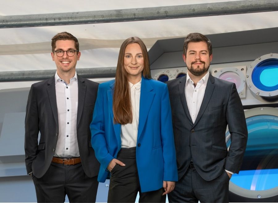Drei Personen in Businesskleidung stehen lächelnd vor einem technischen Gerät mit blauer Optik.