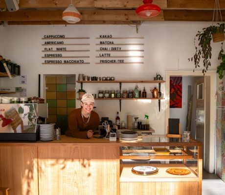 Person hinter Café-Tresen mit Espressomaschine; Wandmenü zeigt Getränke wie Cappuccino, Matcha, Latte; Glasvitrine mit Gebäck.
