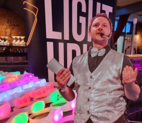 Mann mit Headset-Mikrofon und Fliege präsentiert bunte leuchtende Objekte vor einem „Light Up"-Banner.