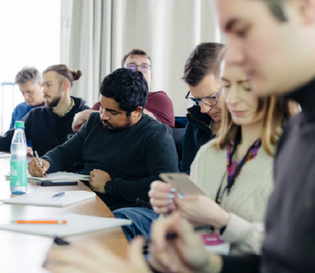 Mehrere Personen sitzen an einem Tisch, schreiben und schauen auf Smartphones. Eine Frau trägt ein Lanyard.