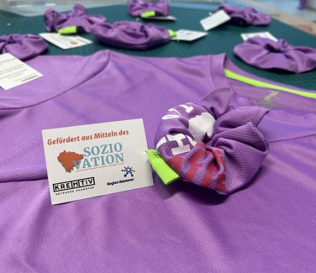 Lila T-Shirts auf grüner Unterlage mit Scrunchies und Karte „Gefördert aus Mitteln des Sozio Nation, Region Hannover".