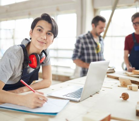 Auszubildende mit Stift und Zettel, Notebook auf der Werkbank. Im Hintergrund der Ausbilder und ein Kollege.
