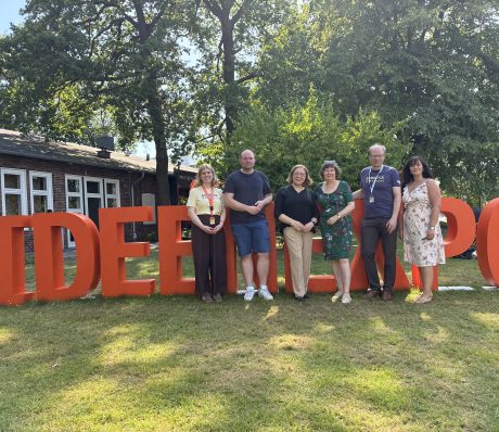 Sechs Personen stehen vor großen orangefarbenen Buchstaben „IDEE EXPO" auf einer Rasenfläche bei Sonnenschein.