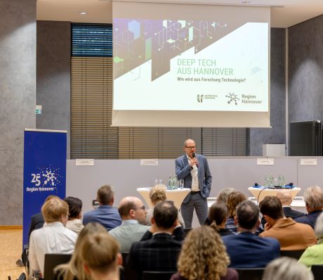 Mann präsentiert mit Mikrofon vor Publikum. Im Hintergrund Präsentationsfolie Deep Tech Hannover.