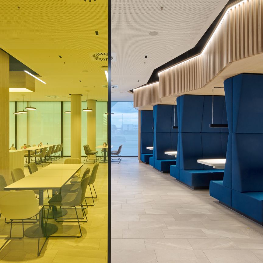 Modernes Büro: links gelb verglaster Meetingraum mit Tisch und Stühlen, rechts blaue Sitzkojen mit weißen Tischen.