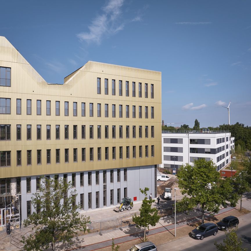 Modernes Bürogebäude mit goldfarbener Fassade im Bau, dahinter weißes Gebäude, Windrad und Bäume unter blauem Himmel.
