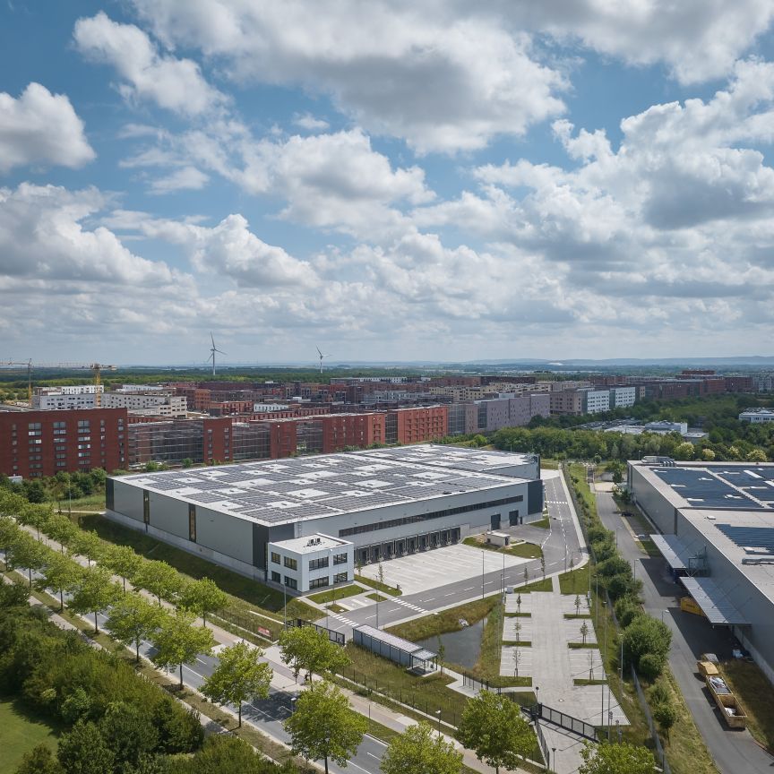 Luftaufnahme eines Industriegebiets mit Lagerhallen und Solardächern, angrenzend an Wohngebäude und Grünflächen.