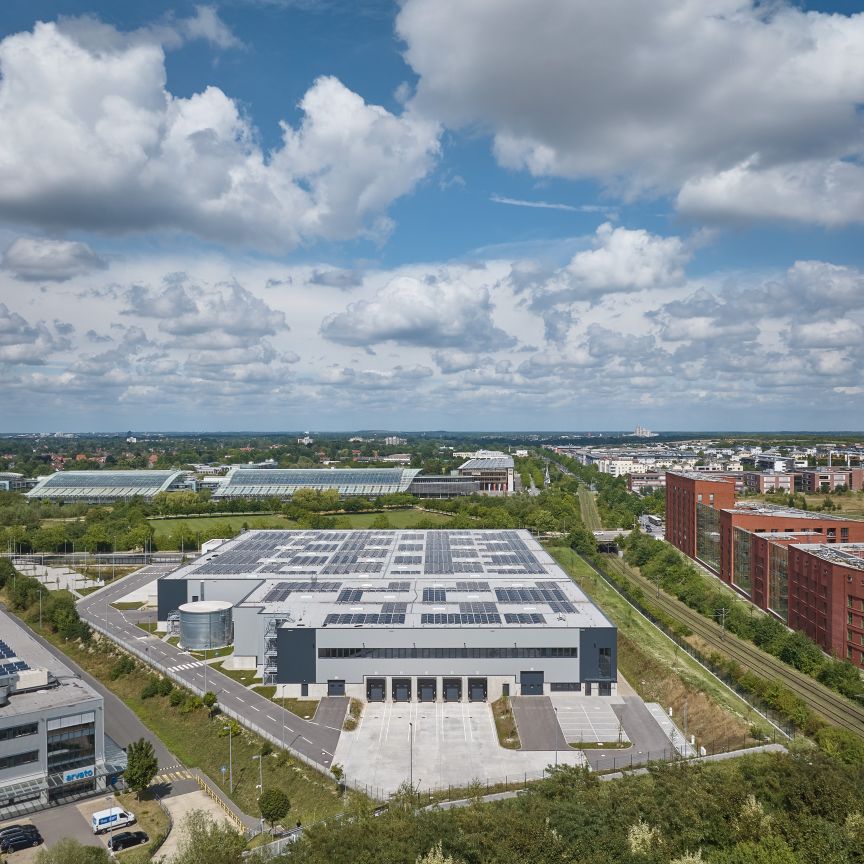 Luftaufnahme eines Industriegebiets mit Lagerhalle, Solarpanelen auf Dächern, Wohngebäuden und Grünflächen unter bewölktem Himmel.