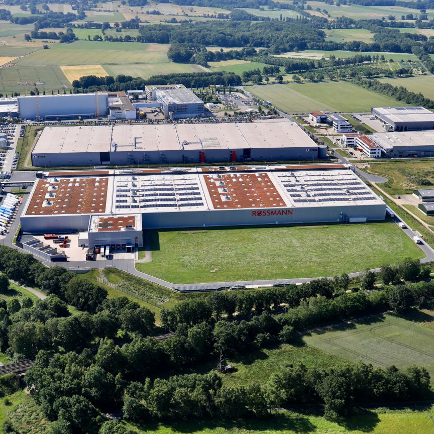 Luftaufnahme eines großen Industriekomplexes mit mehreren Hallen, umgeben von Feldern, Bäumen und einer Autobahn.