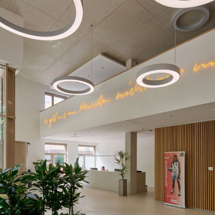 Modernes Foyer mit runden Deckenleuchten, Neonschrift auf Deutsch, Holzwandverkleidung, Pflanzen und großen Fenstern.