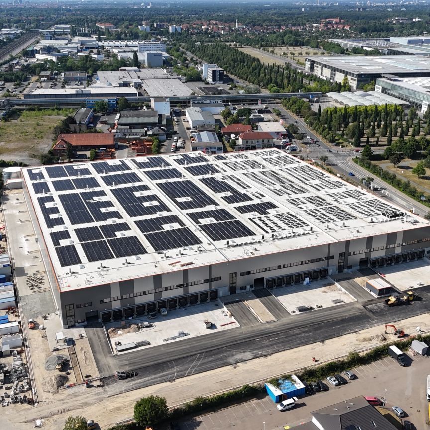 Luftaufnahme eines großen Industriegebäudes mit Solaranlage auf dem Flachdach, umgeben von Industriegebiet und Grünflächen.