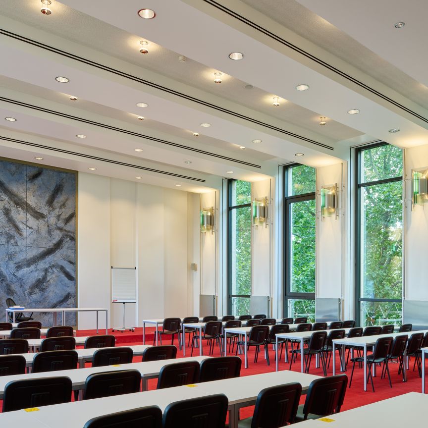 Konferenzraum mit Stuhlreihen, rotem Teppich, großen Fensterfronten, Wandgemälde und Deckenbeleuchtung.