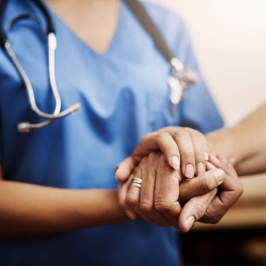 Pflegeperson in blauen Scrubs mit Stethoskop hält beidseitig die Hände einer anderen Person.