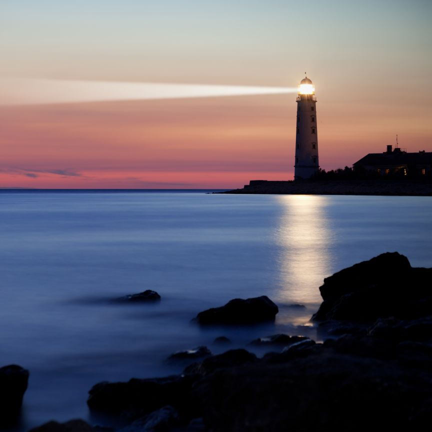 Leuchtturm an felsiger Küste bei Dämmerung, Lichtstrahl über ruhiges Wasser, rosa-blauer Abendhimmel.