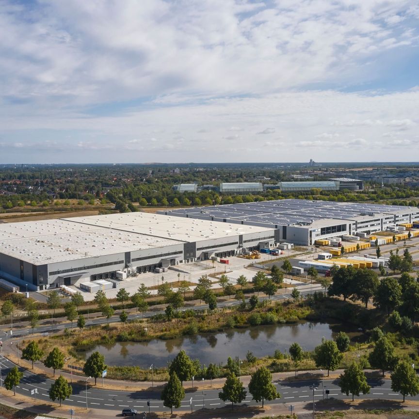 Luftaufnahme eines großen Industriekomplexes mit Lagerhalle, Solaranlage, Parkplatz und Teich, umgeben von Bäumen.