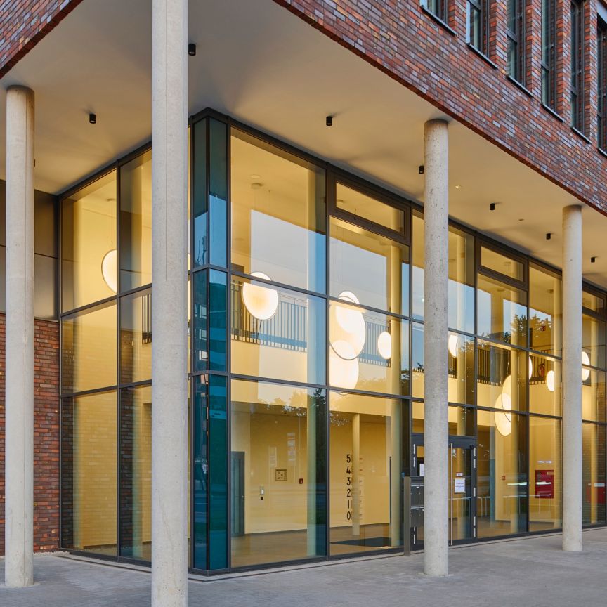 Modernes Bürogebäude mit roter Backsteinfassade, großen Glasfronten, runden Deckenleuchten und Betonsäulen im Eingangsbereich.