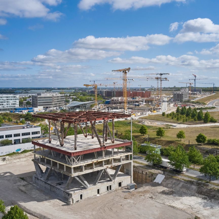 Luftaufnahme einer Großbaustelle mit mehreren Turmdrehkranen, einem Rohbau im Vordergrund und Bürogebäuden im Hintergrund.