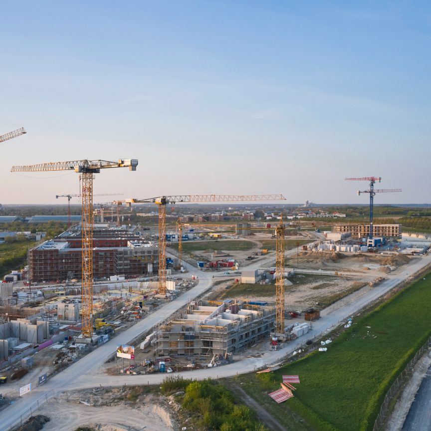 Luftaufnahme einer Großbaustelle mit mehreren Turmkränen, teilfertigen Gebäuden, Baustraßen und umliegenden Grünflächen.