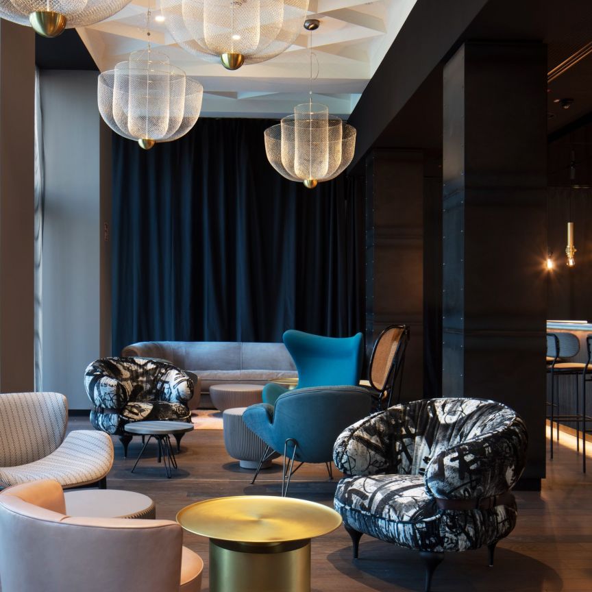 Moderne Hotellobby mit gemusterten Sesseln, blauem Sofa, goldenen Beistelltischen, großen Hängelampen und einer Bar im Hintergrund.