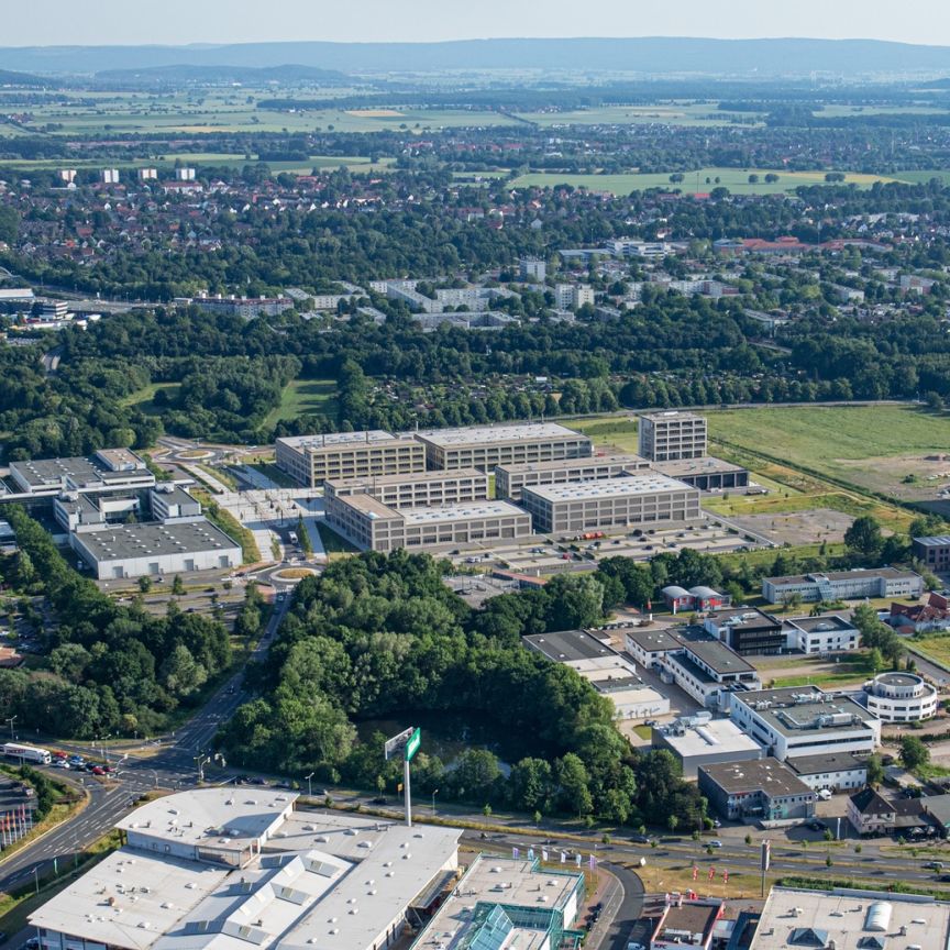 Luftaufnahme einer Stadt mit Gewerbegebiet, Bürogebäuden, Grünflächen und Feldern im Hintergrund bei sonnigem Wetter.