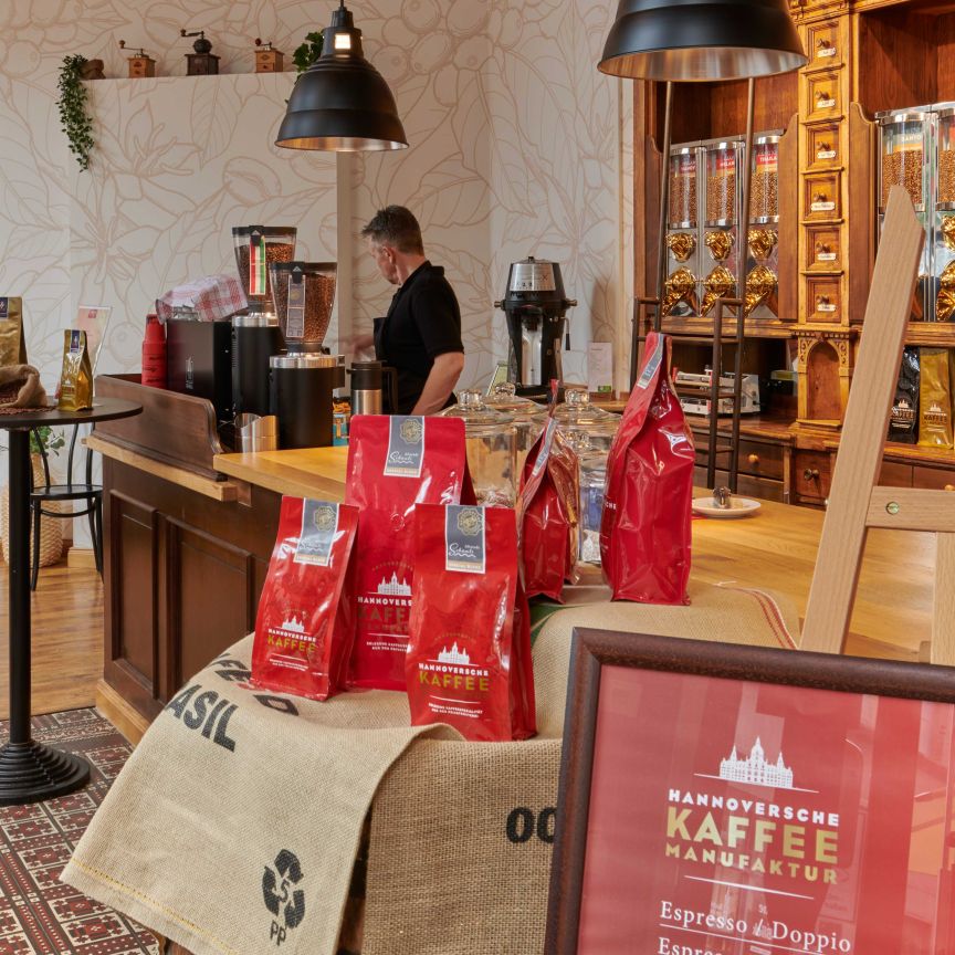 Kaffeeshop der Hannoverschen Kaffeemanufaktur mit roten Kaffeepaketen, Holztheke, Kaffeebohnen-Schrank und einem Mitarbeiter.