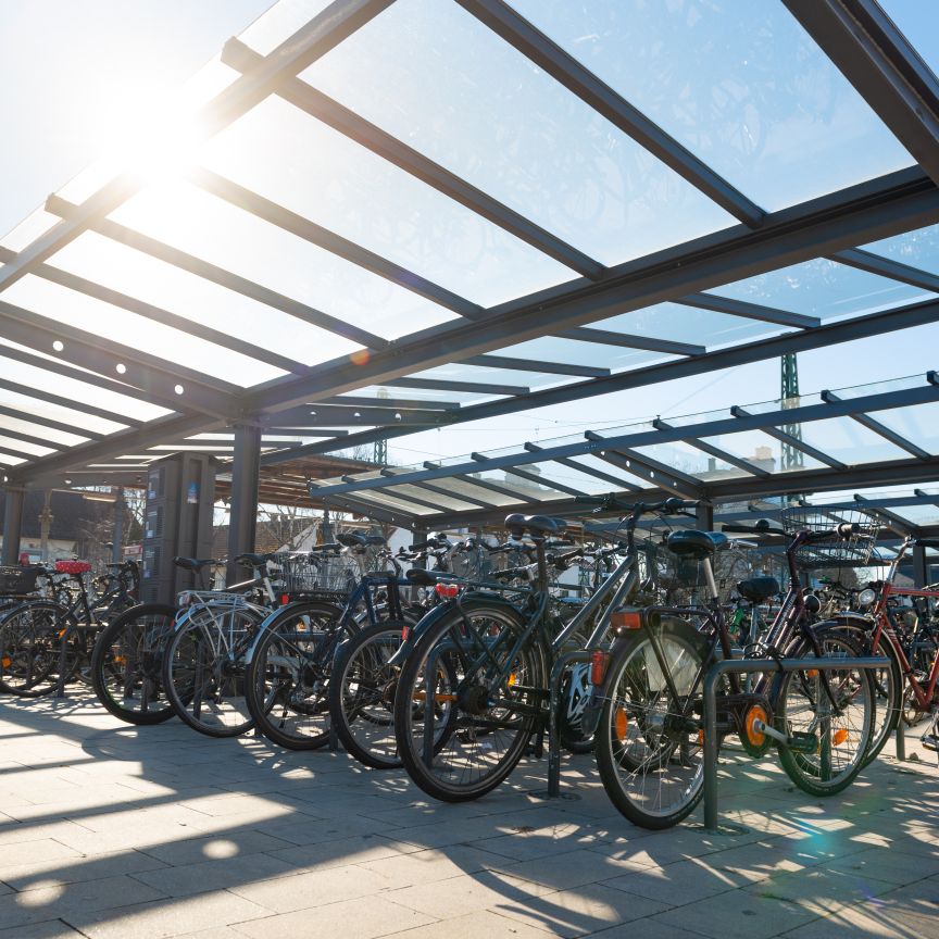 Überdachter Fahrradparkplatz mit zahlreichen abgestellten Fahrrädern unter einem Stahl-Glas-Dach bei Sonnenschein.