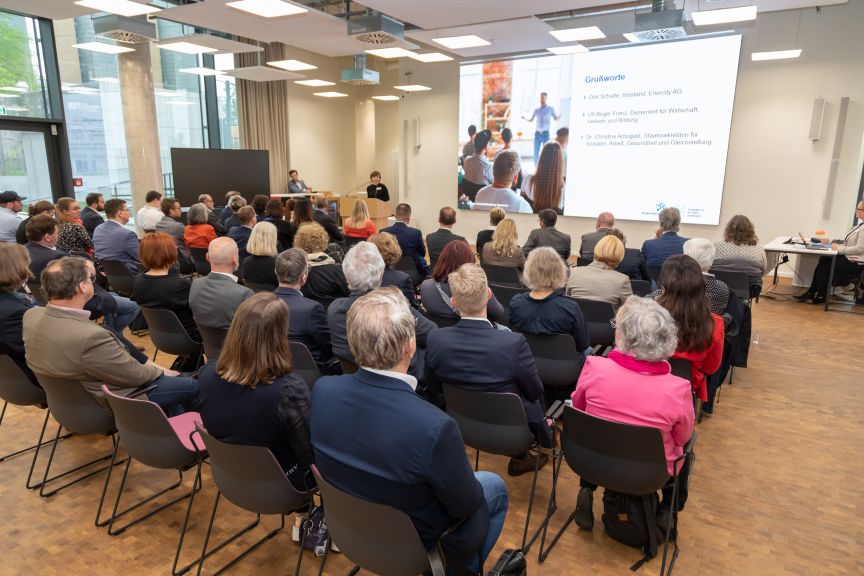 Publikum in einem Konferenzraum blickt auf eine Leinwand mit deutscher Präsentation; vorne steht eine Rednerin am Podium.