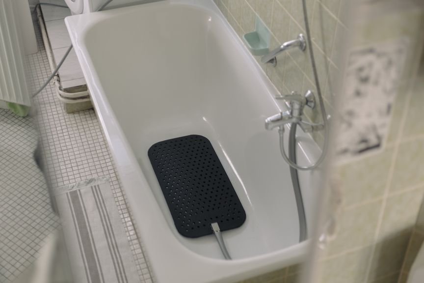 Weiße Badewanne mit schwarzer Gummimatte und Kabel im Inneren, Duscharmatur, Kacheln, Badezimmer.