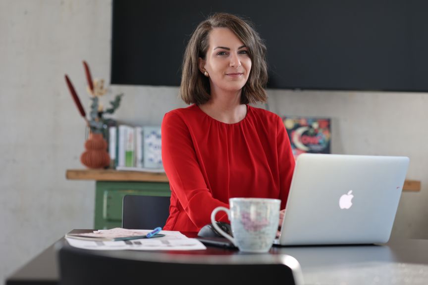 Frau in roter Bluse sitzt an einem Tisch mit Apple-Laptop und Tasse, im Hintergrund ein Regal.