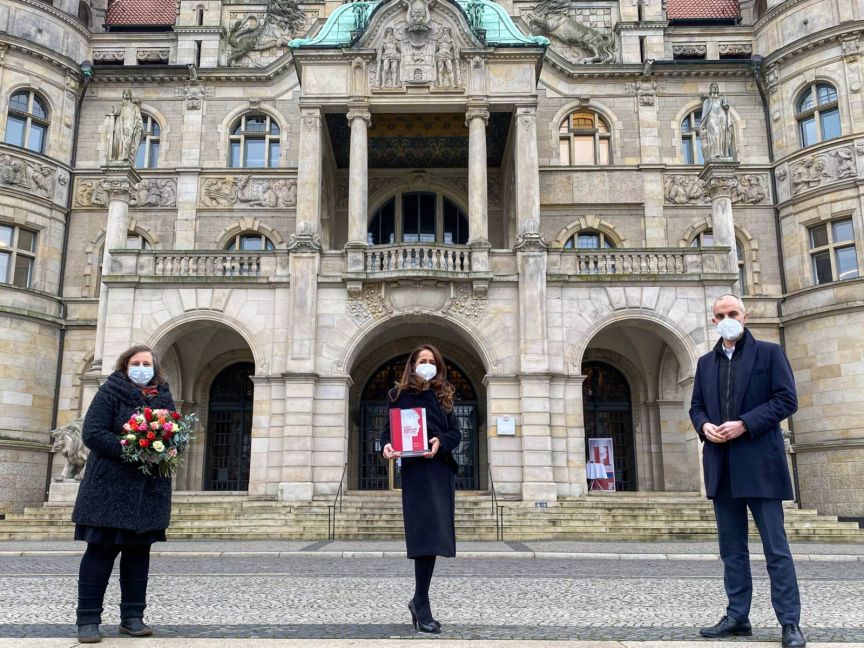 Drei Personen mit Masken stehen vor einem historischen Gebäude. Eine hält Blumen, eine andere ein Buch/Heft.