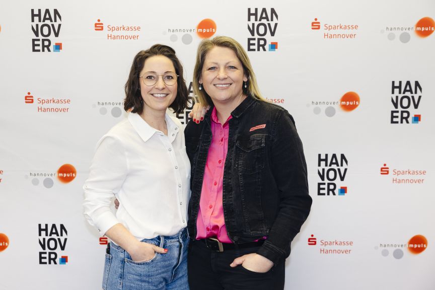 Zwei Frauen stehen lächelnd vor einer Fotowand mit Logos von Sparkasse Hannover und Hannover Impuls.
