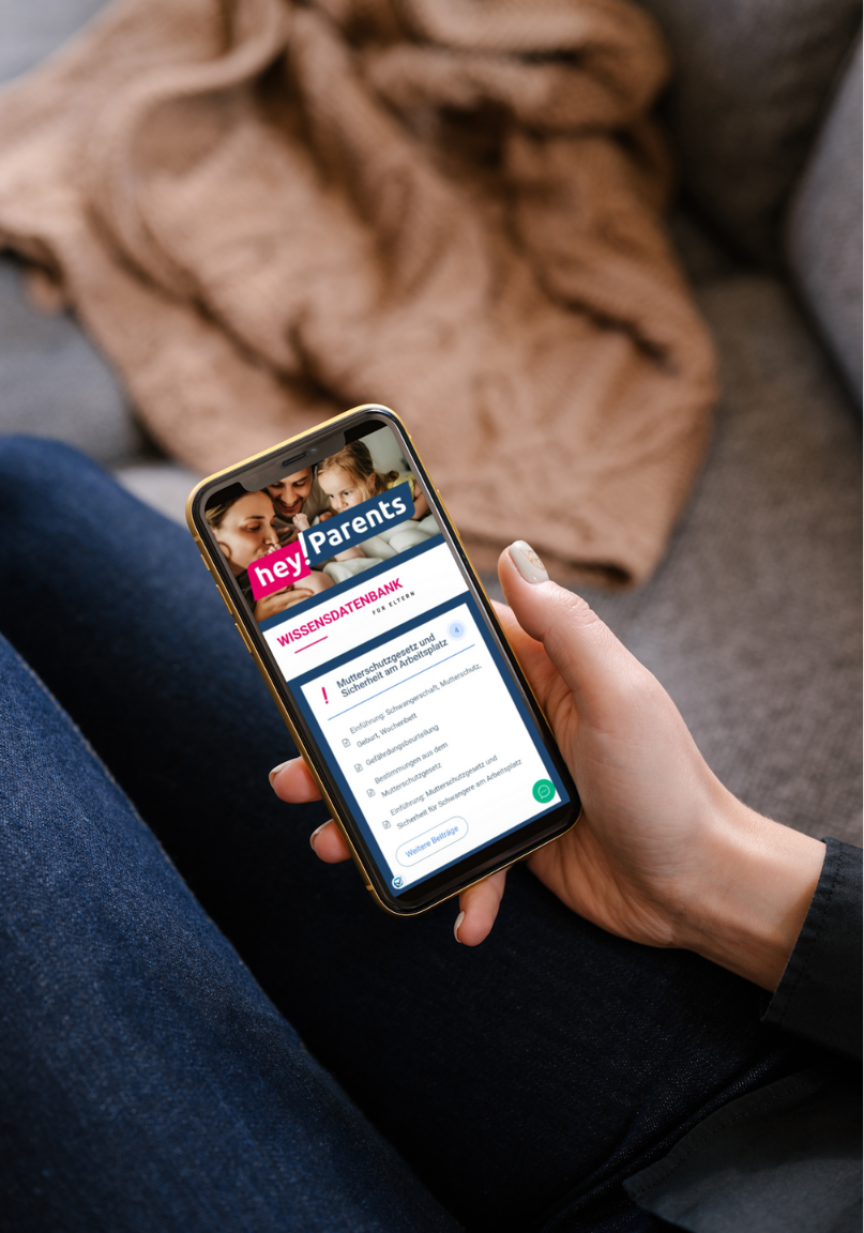 Person hält Smartphone mit geöffneter „hey!Parents"-App, Bereich „Wissensdatenbank", auf einem Sofa sitzend.