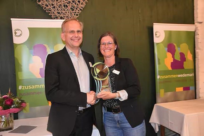 Zwei Personen halten gemeinsam eine Trophäe. Im Hintergrund Werbebanner mit Hashtag #zusammen.