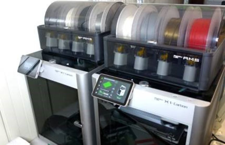 Zwei Bambu Lab X1 Carbon 3D-Drucker mit AMS-Einheiten und mehreren Filamentspu­len, daneben ein Tablet-Display.